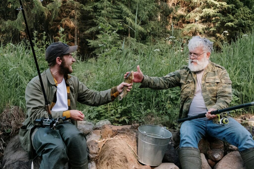 Two men share a moment while fishing in a lush, green countryside area.